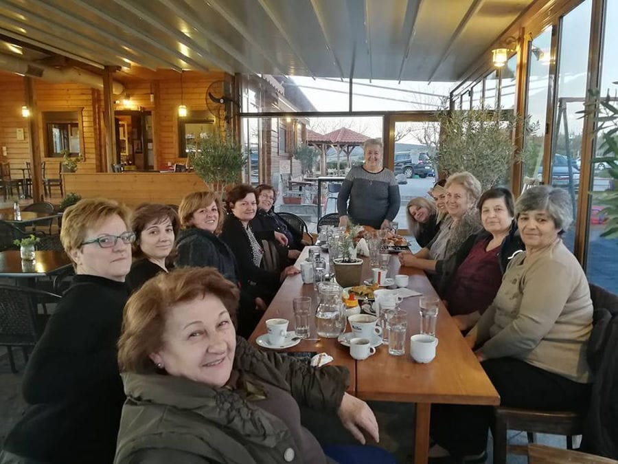 a group of women around the table at 'Agios Antonios Women’s Agri Cooperative' stone building and watching at the camera